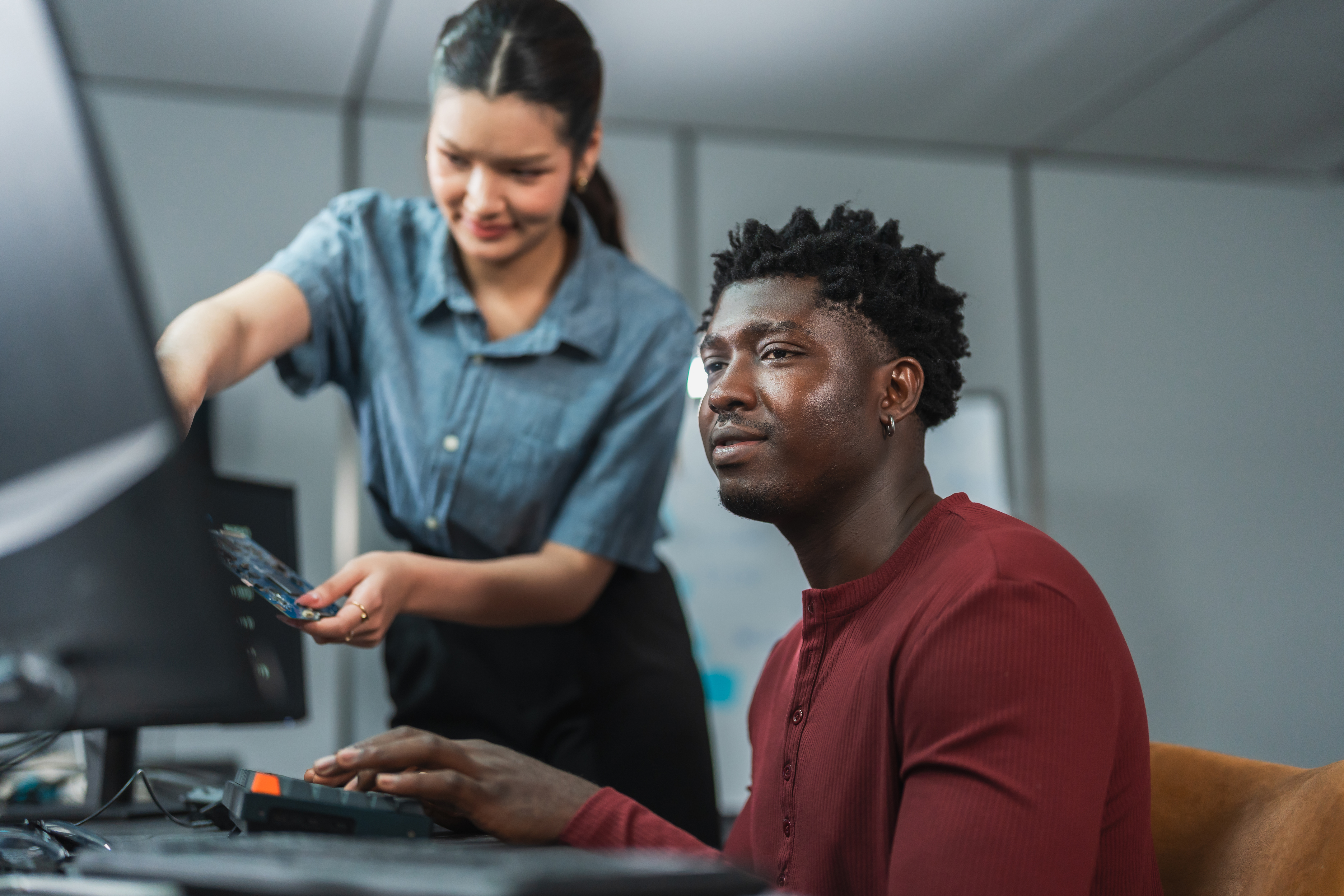 Asian engineer holds a circuit board while an African American developer looks at it from his desk in a technology office.